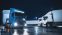 Blue Semi-Truck with Cargo Trailer Drives Off From Overnight Parking Space where Other Trucks are Standing. Long Haul Truck Leaves Parking Lot, Transporting Cargo / Goods Across Continent. Rainy Night